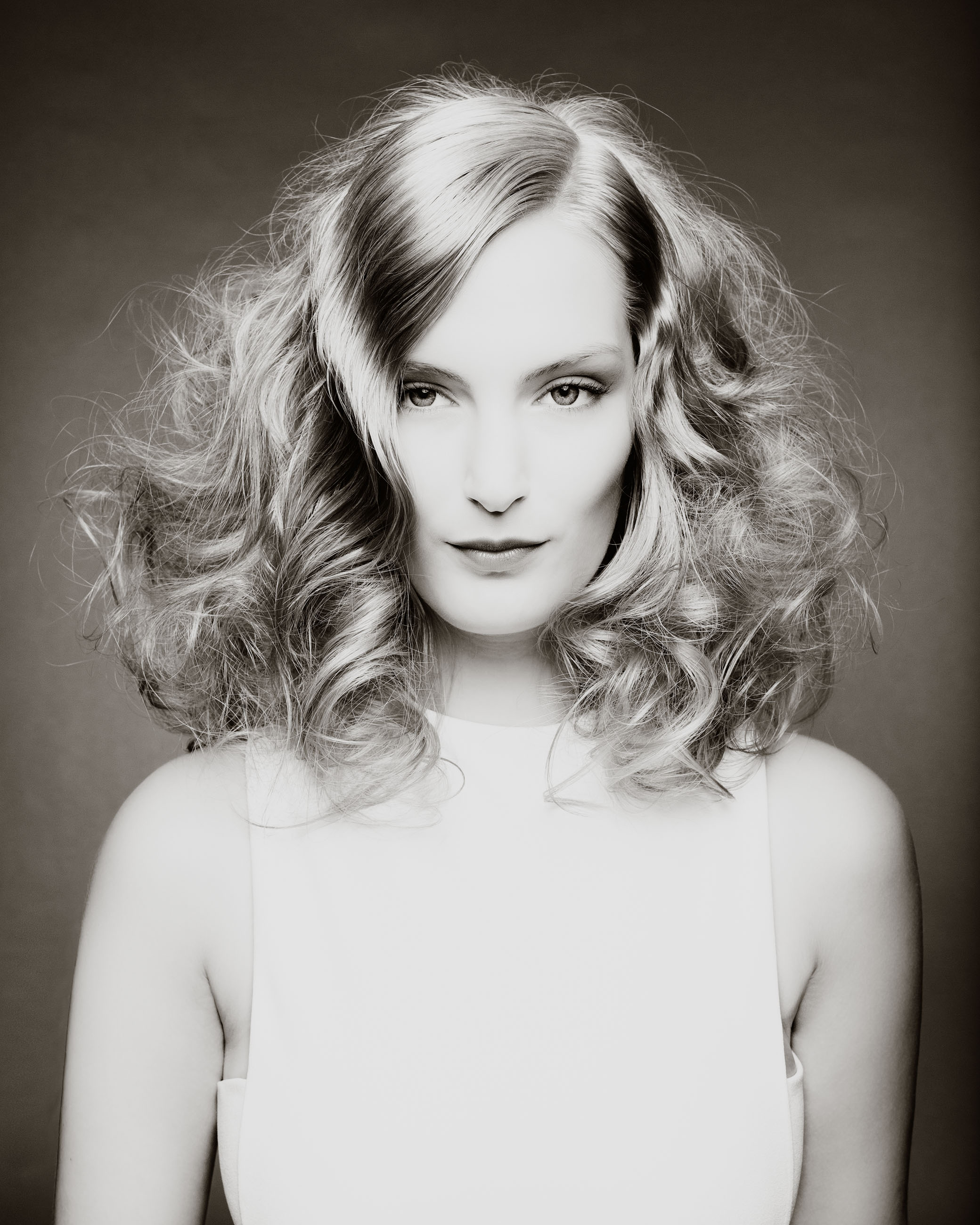 Portrait Blonde Frau mit lockigen Haaren und hellem Makeup vor grauem Hintergrund Schwarz Weiß Sven Vogel Photograph Fotograf